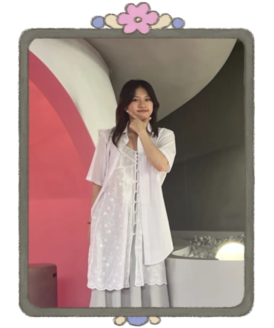 Emily smiling with her hand on her chin, wearing a white lace blouse in front of a pink arch